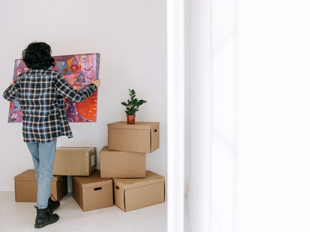 A Decluttering Guide for a Stress-Free Move 3 A person stands near stacked cardboard boxes and a potted plant, holding a colorful painting against a white wall—perhaps arranging or using a Decluttering Guide to organize and decorate their space.