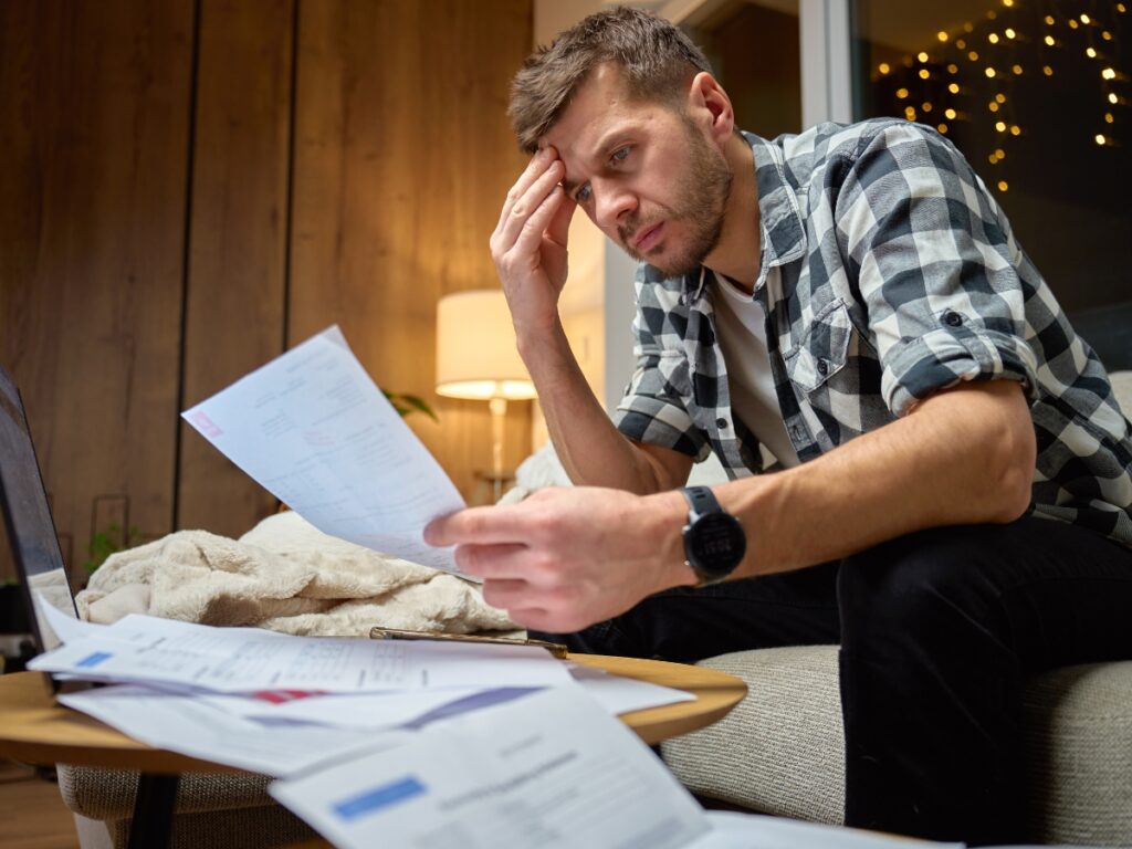 Utility Disconnection Guide Before Moving 1 A man sits on a couch reviewing bills, looking concerned about potential utility disconnection, with paperwork spread out on the table in front of him.