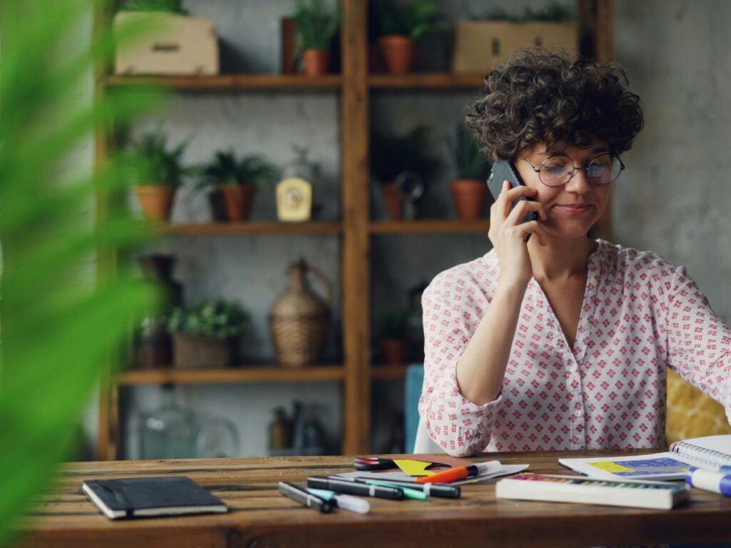 Utility Disconnection Guide Before Moving 3 A person with curly hair and glasses sits at a wooden table, talking on the phone about a utility disconnection, surrounded by notebooks and pens, with plants on shelves in the background.